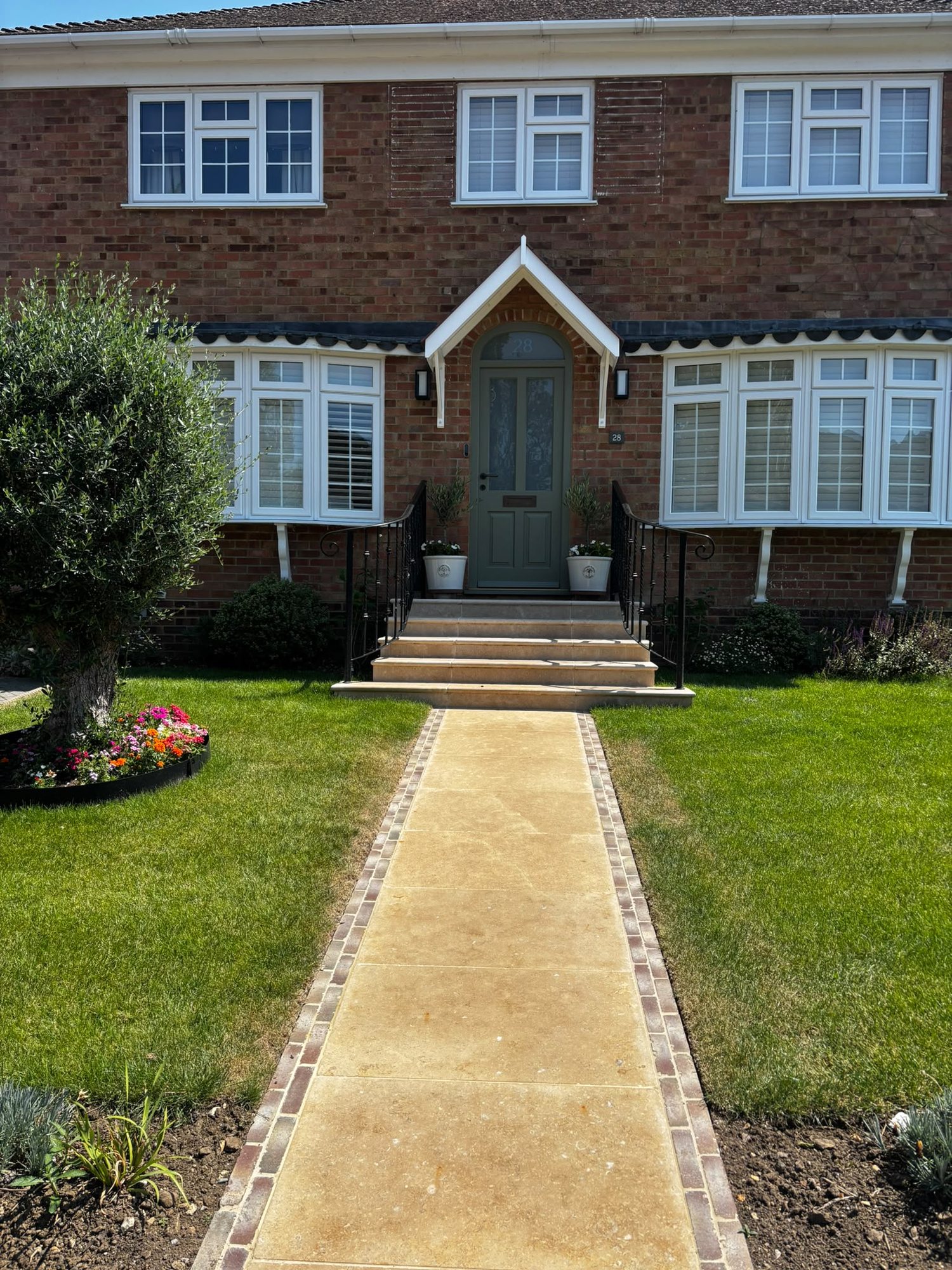 The same house after the work: a sage green front door under a new pitched porch canopy with a brick arch over it, new limestone steps and path with a brick soldier-course edge, planters either side of the door and a level lawn