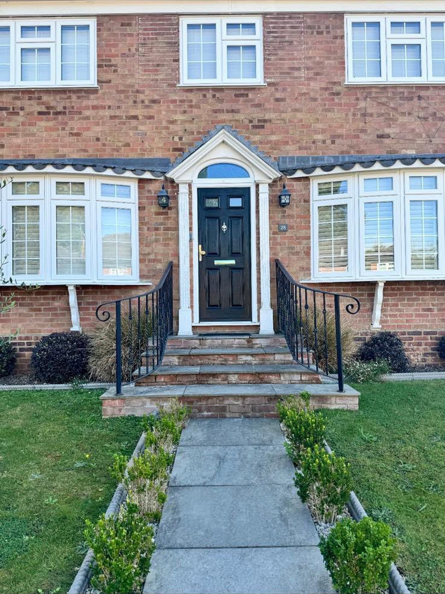 The front of a red brick house before the work: a dark front door under a white columned porch with a triangular pediment, worn grey concrete steps and a cracked slab path between thin borders