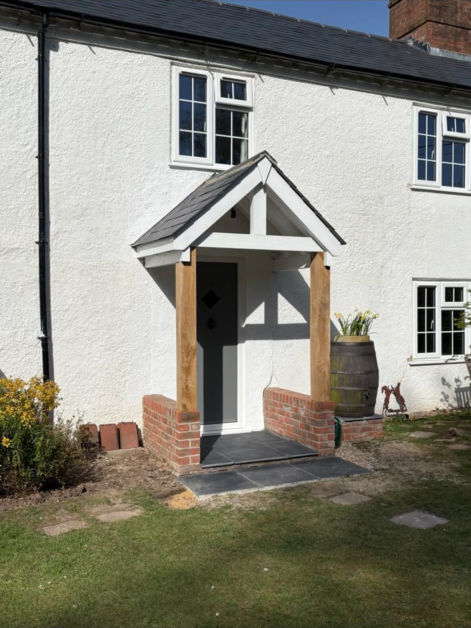The same cottage after the work, with a new green oak porch on red brick dwarf walls, a pitched slate roof, a slate floor and a new grey front door