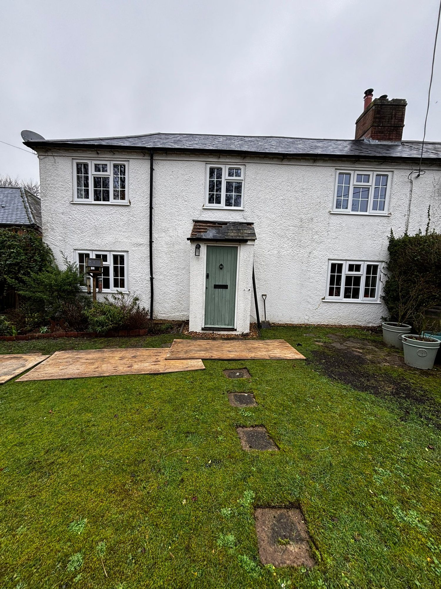 A white rendered cottage before the work, with a small shallow-pitched porch in poor condition over a green plank door, and boards laid across the lawn to protect it